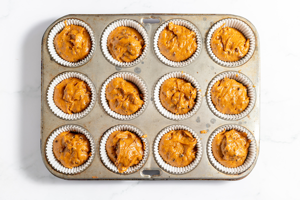 Image, taken from above, shows a muffin tin with the batter for pumpkin muffins poured into each cup, ready to be baked.
