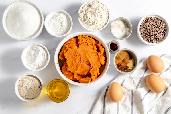 Image, taken from above, shows the ingredients needed to make gluten free pumpkin muffins, incluging pumpkin puree, various gluten free flours, eggs, oil, and seasonings.