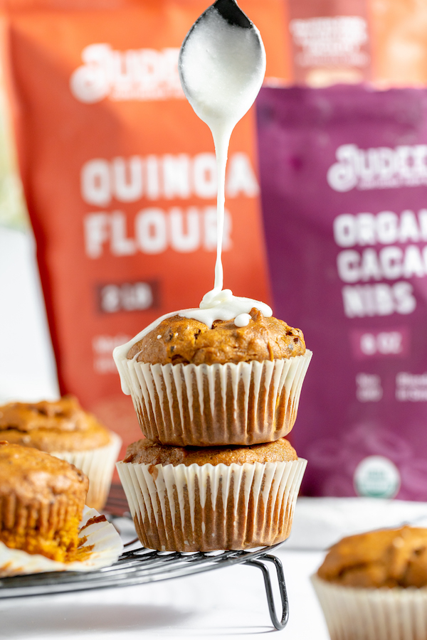 Image shows two pumpkin muffins stacked on one another, with vanilla glaze being drizzled onto them. Judee's products sit in the background of the photo.