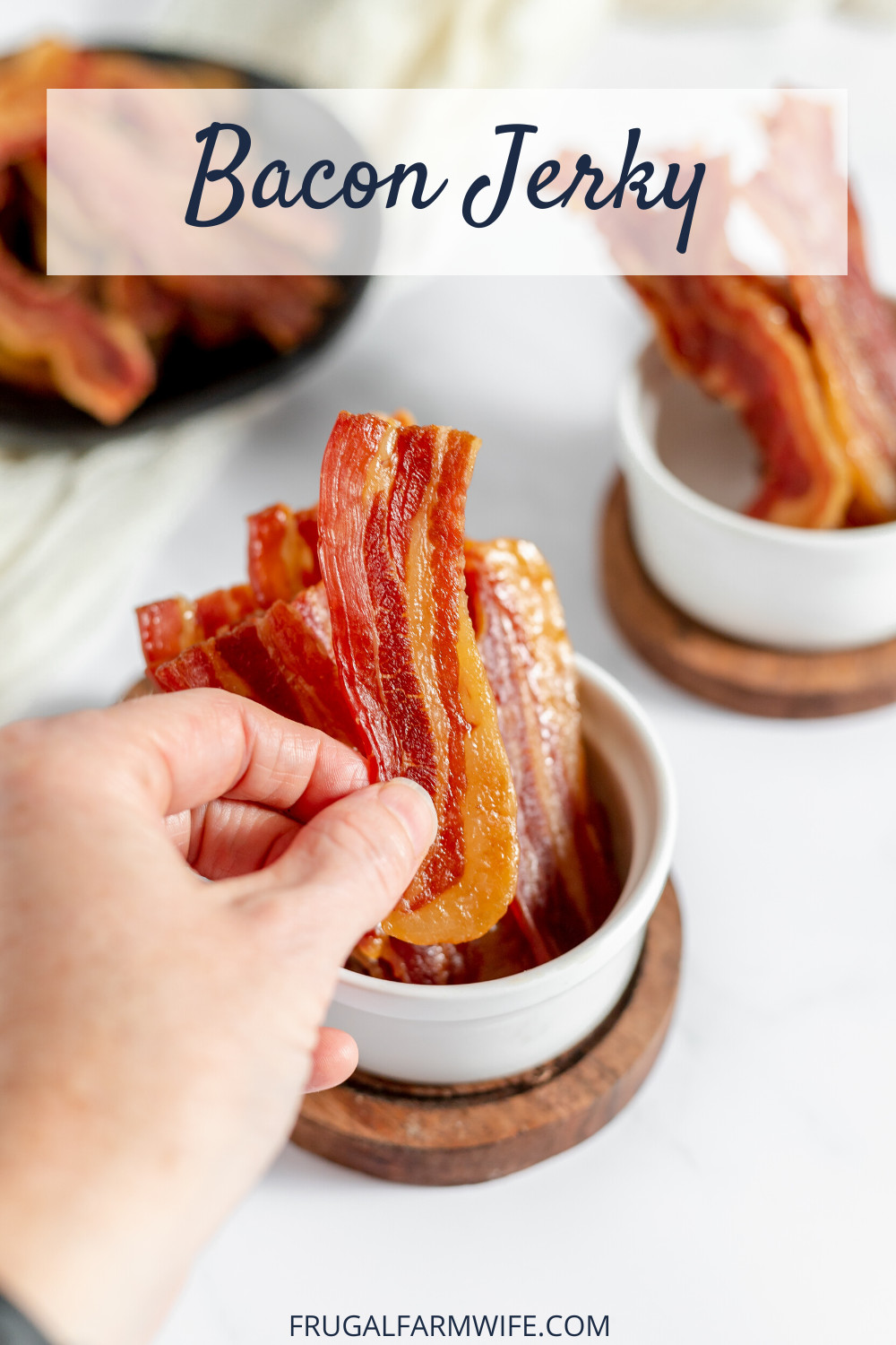 Image shows a white table with small ramekins of bacon jerky. The front ramekin has a hand holding a slice of bacon jerky. Text above reads "Bacon Jerky"