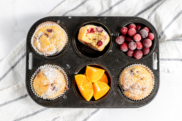 Image shows a small muffin tin with three gluten free cranberry orange muffins, one tin holds cranberries, one holds orange slices, and the last holds a partially eaten muffin.