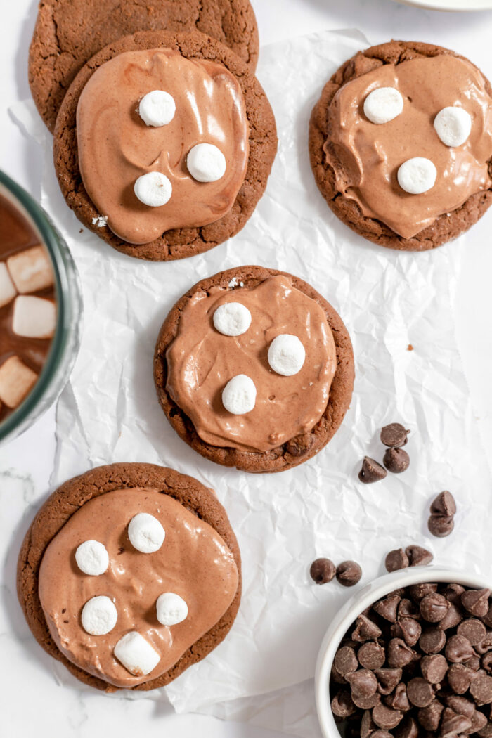Image, taken from above, shows several hot chocolate cookies on a parchment paper, with small marshmallows on each. 