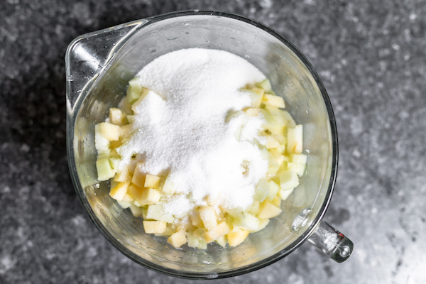 Image, taken from above, shows a large glass measuring cup full of small sliced apples and sugar on top