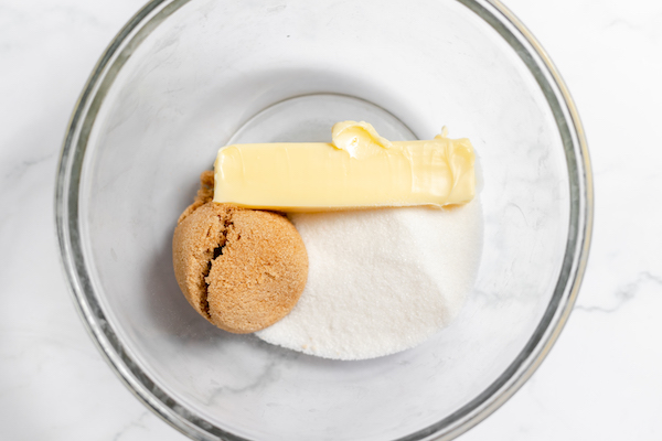Image, taken from above, shows softened butter, sugar and brown sugar in a glass mixing bowl. 