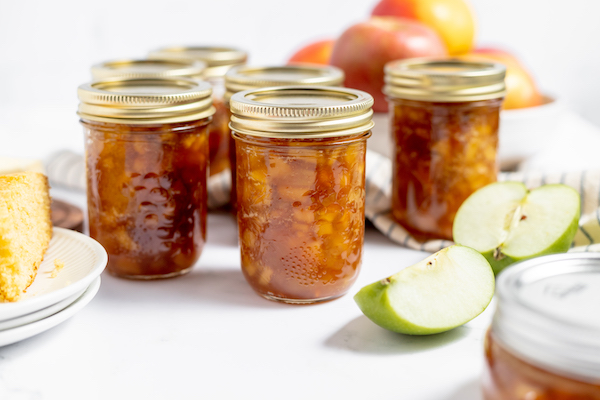 Image shows several jars of apple jam on a white table, with a green apple sliced in half sitting nearby. In the background is a bowl of red apples in a white bowl