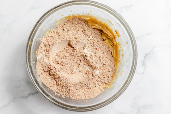 Photo, taken from above, shows dry ingredients for hot chocolate cookies sprinkled on top of the batter for the cookies in a glass bowl. 