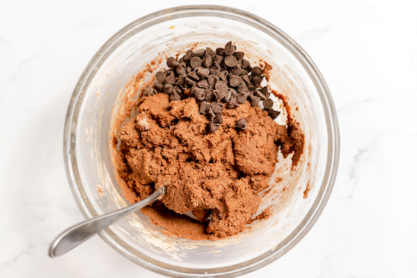 Photo shows hot chocolate cookie batter in a glass mixing bowl, with chocolate chips sprinkled on top. 