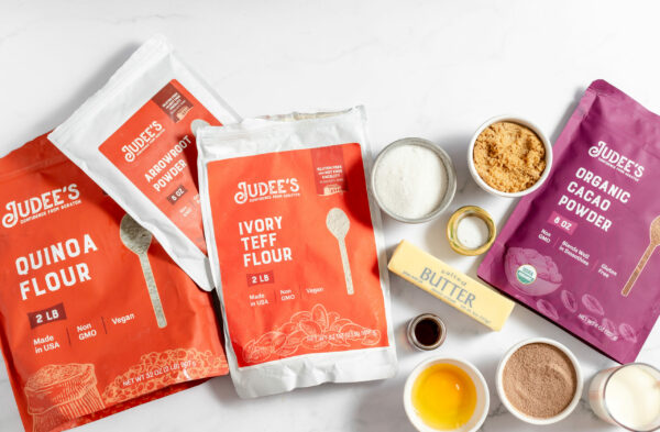 Image shows a variety of Judee's flour products on a counter, along with other ingredients to make gluten free hot chocolate cookies- sugar, brown sugar, vanilla, butter, and more. 