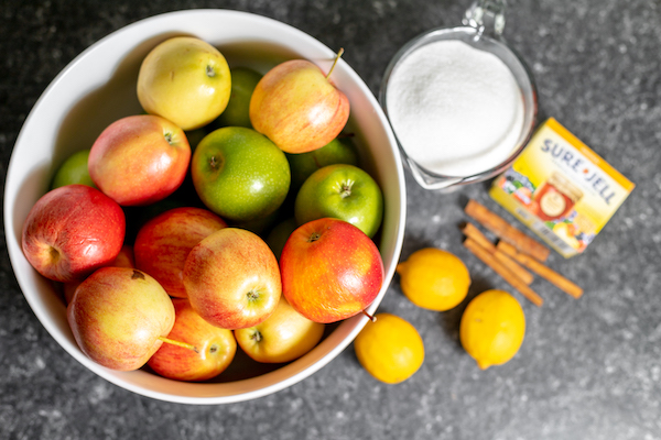 Image taken from above shows a large white bowl full of apples, several lemons, sticks of cinnamon, a measuring cup of sugar, and a package of gelatin on a counter
