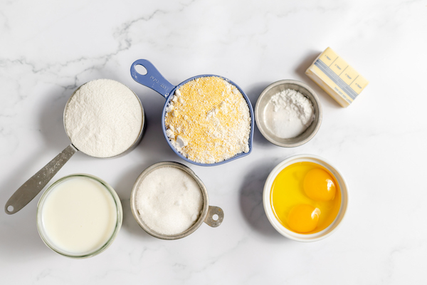 Image, taken from above, shows ingredients needed to make gluten free corn bread- including cornmeal, rice flour, quinoa flour, arrowroot powder, sugar, baking powder, salt, eggs, milk and melted butter. 
