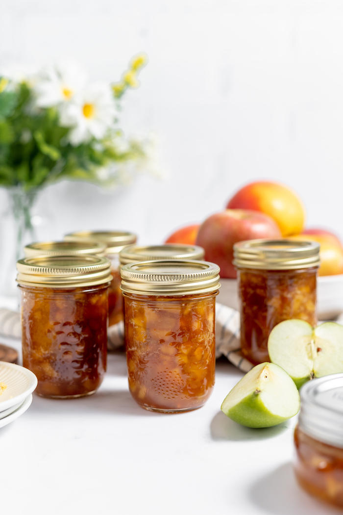 Photo shows several jars of apple jam on a white table surrounded by apple slices and a white and blue towel