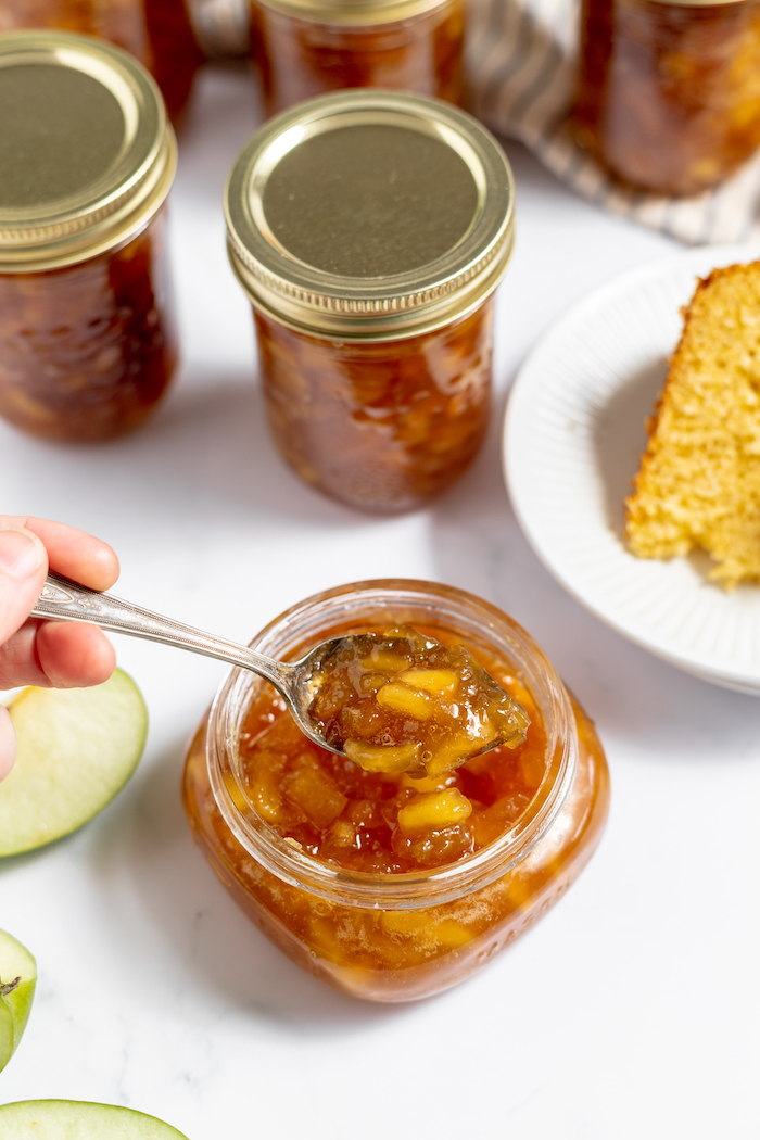 Image shows a woman scooping a spoonful of homamde apple jam from a small glass jar. The table is filled with larger sealed jars of hDIY apple jam