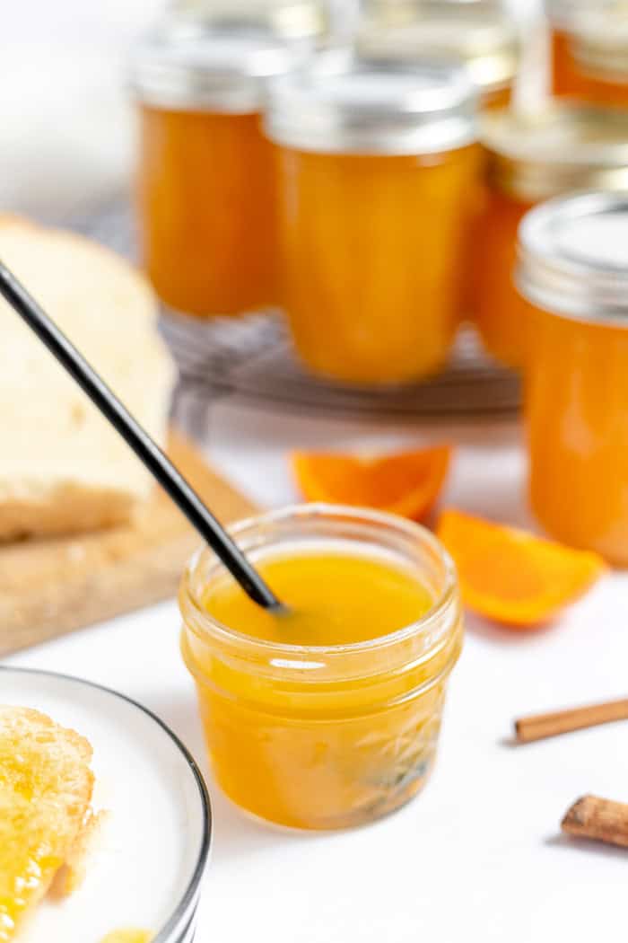 Image shows a small jar of orange jam with a black spoon sticking out. In the background are more jars of orange jam sitting on a cooling rack.