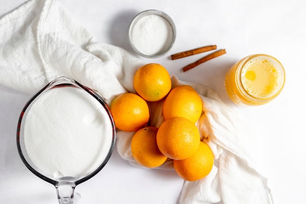 Image, taken from above, shows a glass measuring cup of sugar, pile of oranges, jar of orange juice and sticks of cinnamon.