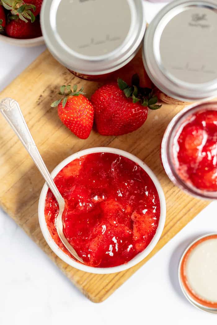 Image, taken from above, shows a small bowl of strawberry jame with a spoon in it. It sits on a wooden cutting board next to a jar lid, some fresh strawberries, and two jars of jam. 