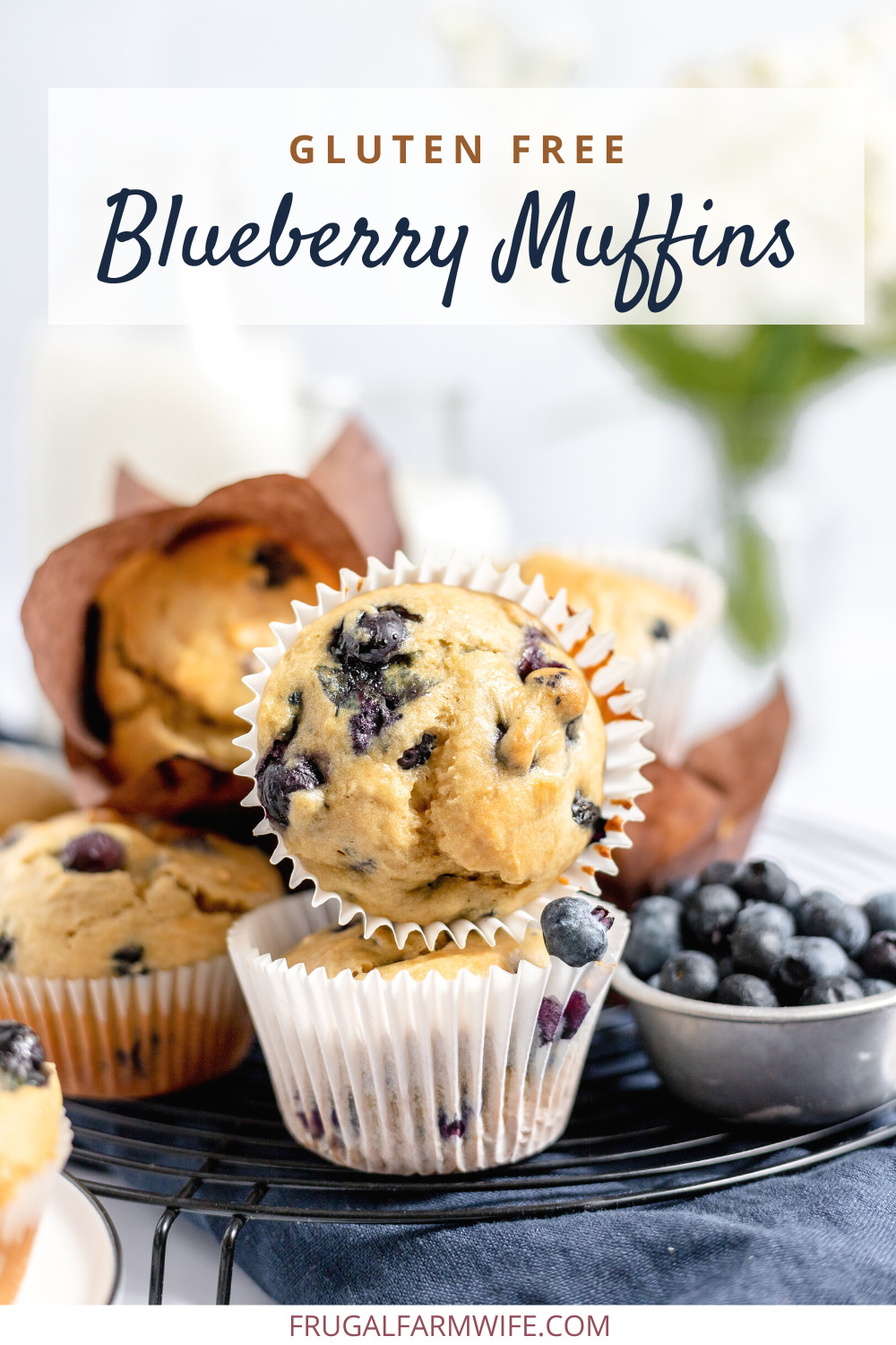Image shows several gluten free blueberry muffins piled on one another on a wire rack. Nearby is a small bowl of fresh blueberries. Text above reads "Gluten Free Blueberry Muffins"