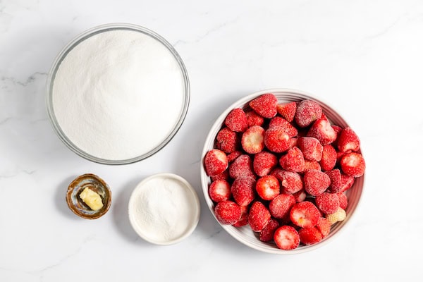 Image, taken from above, shows several bowls on a white counter with the ingredients to make strawberry jam. A bowl of sugar, frozen strawberries, pectin, and some butter.