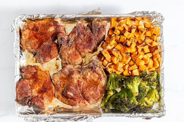 Image, taken from above, shows a sheet pan covered in foil with roasted pork chops, sweet potatoes and broccoli. 