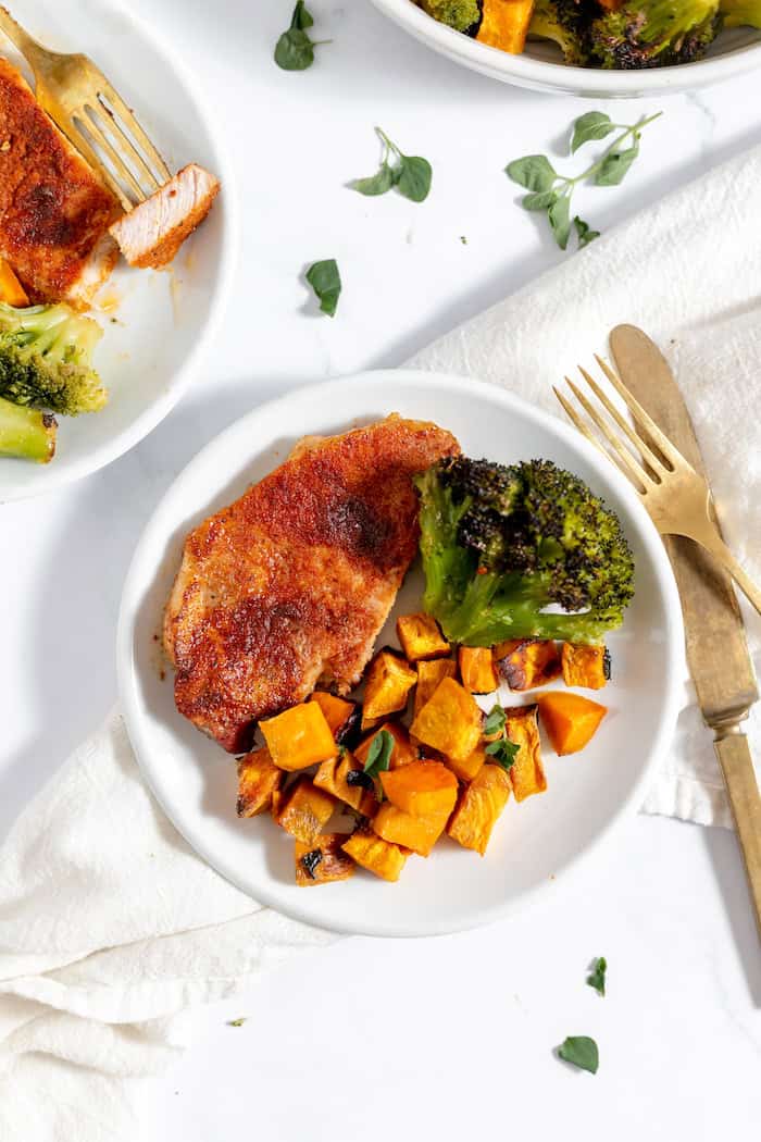 Image shows a plate with a serving of pork chops, roasted sweet potatoes and broccoli on a white table cloth. Next to the plate sits a fork and knife. 