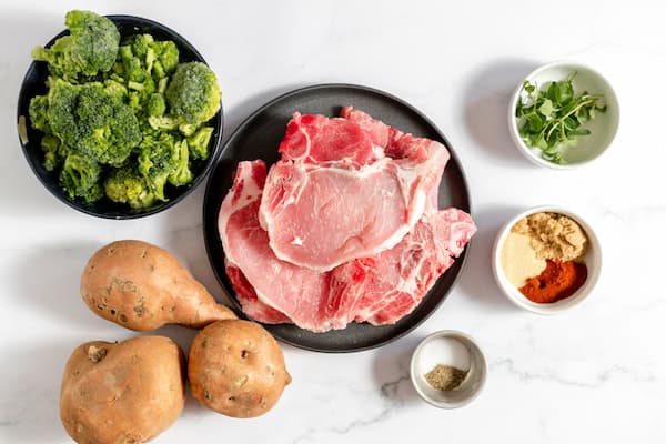 Image, taken from above, shows the ingredients for this one pan meal- broccoli in a bowl, a plate of pork, sweet potatoes, herbs and spices.