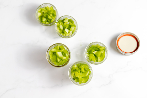 Image, taken from above, shows 5 small mason jars with sliced celery in each. The lid for one jar sits nearby.