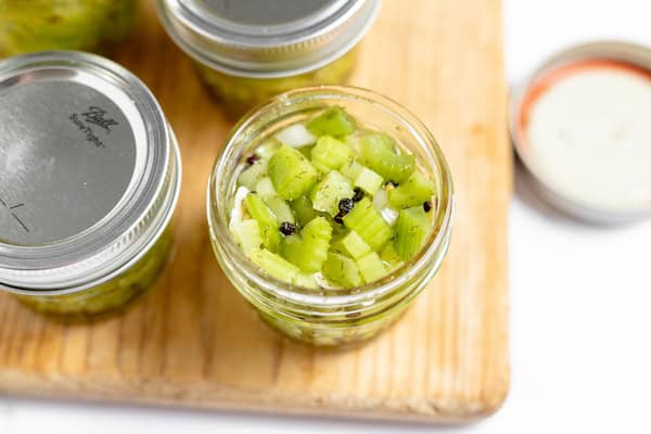 Image, taken from above, shows several jars of diced celery sitting on a cutting board, with pickling brine poured into the jars. 
