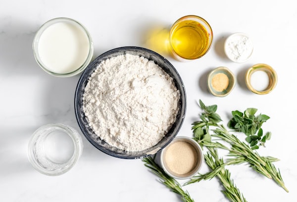 Image, taken from above, shows bowls of flour, milk, oil, salt, yeast, and herbs on a table- all the ingredients for gluten free focaccia bread