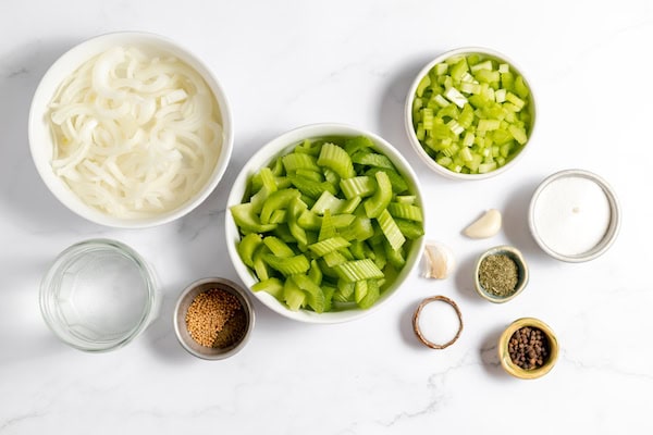 Image, taken from above, shows several bowls laid out on a white surface full of the ingredients needed to pickle celery. Onions, celery, spices and vineager. 