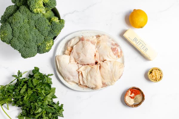Image, taken from above, shows the ingredients for lemon garlic chicken laid out to cook- chicken, broccoli, butter, lemon, and spices.