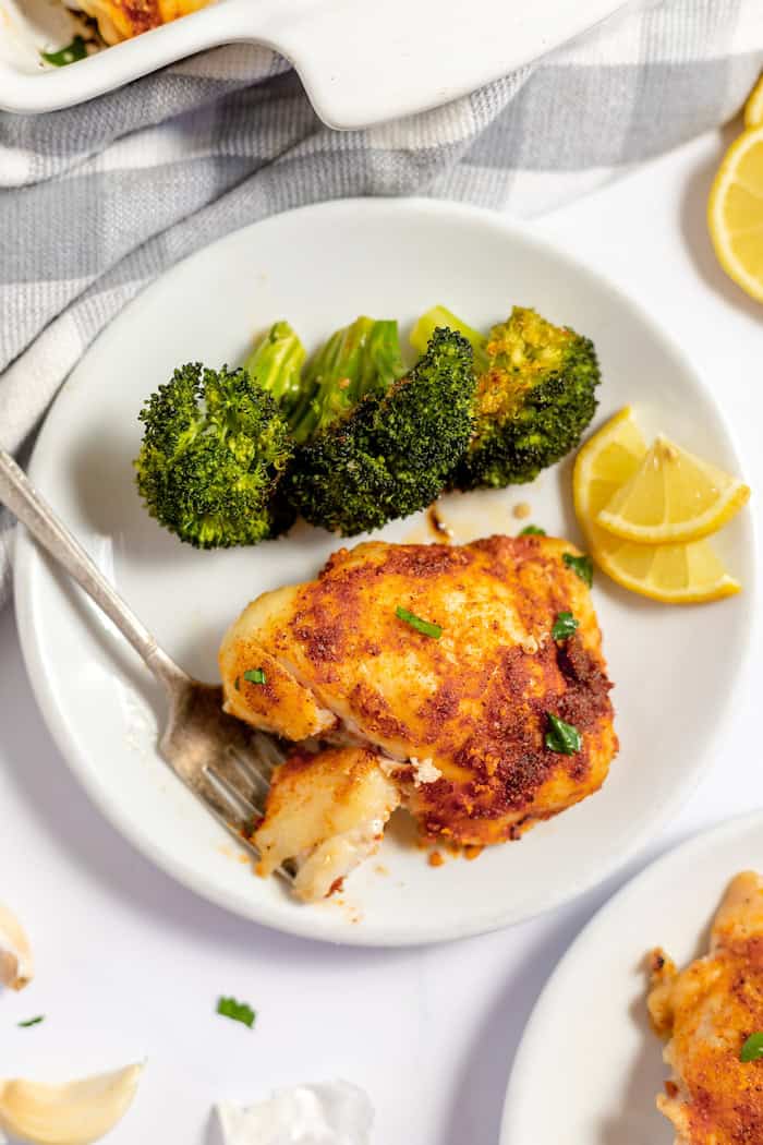 Image shows a white plate with a serving of lemon garlic chicken on it. A fork takes a bite rom the chicken, with some broccoli and lemon slices also on the plate. 