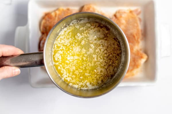 Photo, taken from above, shows melted butter with garlic in it in a saucepan over the casserole dish of prepared chicken thighs