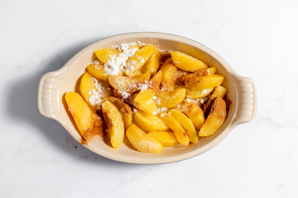 Image, taken from above, shows peach filling topped with seasonings and ready to be topped with the crisp topping. 