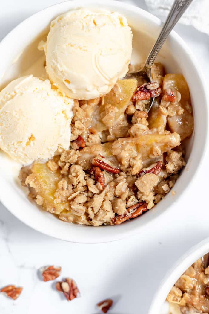 Image, taken from above, shows a serving of gluten free peach crisp in a white bowl, with a spoon and two scoops of vanilla ice cream