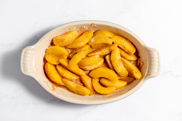 Image, taken from above, shows the peaches mixed in a ceramic dish ready to be topped with crumble topping.