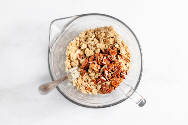Images shows the ingredients for gluten free peach crisp in a glass bowl, with pecans sprinkled on top.