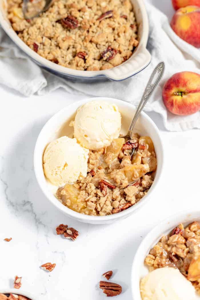 Image shows a small white bowl with a serving of peach crisp and two scoops of ice cream. A larger serving dish of peach crisp sits nearby. 
