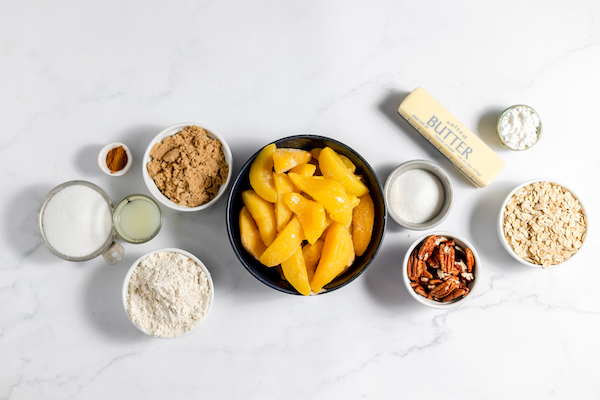 Image, taken from above, shows several small bowls of the ingredients needed to make gluten free peach crisp. Ingredients include sugars, butter, pecans, sliced peaches and more. 