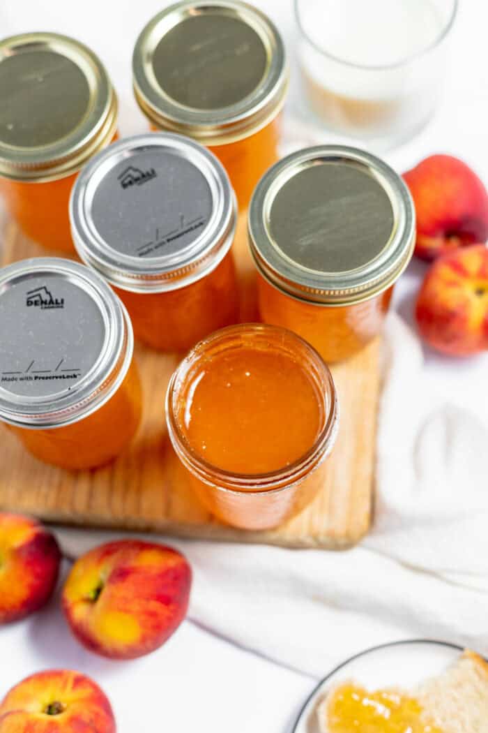 Image shows several canning jars full of peach jam with peaches surrounding them. 