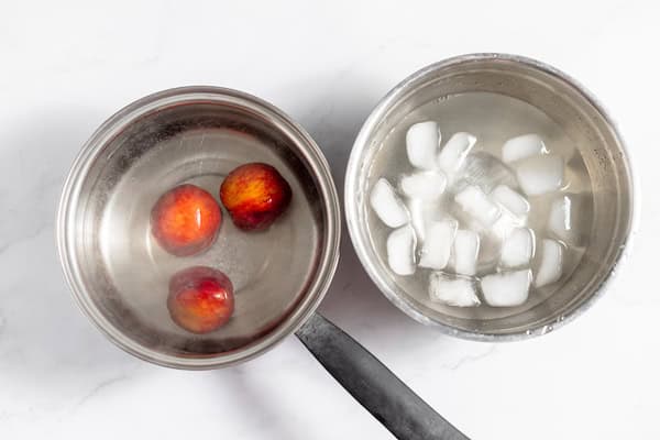 Image, taken from above, shows peaches in a hot water to blanch for peeling. A large pot of ice water sits next to the peaches.