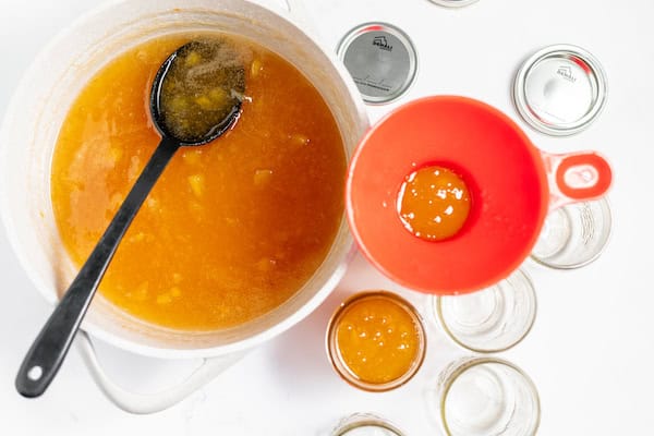Image, taken from above, shows a ladle, spooning fresh peach jam into canning jars.