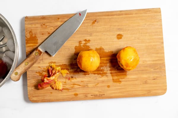 Image, taken from above shows two peaches on a cutting board and a knife peeling skins off of blanched peaches