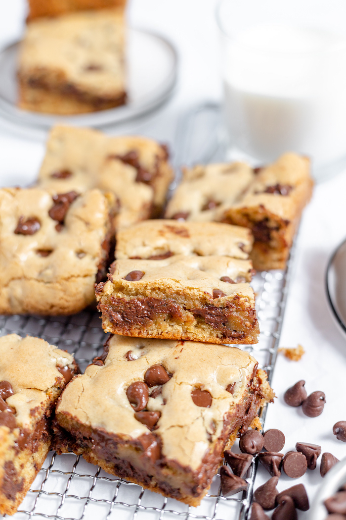 Image shows several gluten free chocolate chip blondies cooling on a cooling rack with chocolate chips scattered nearby. 
