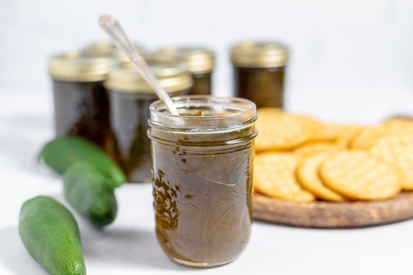Image shows a jar of jalepeno jelly with a spoon sticking out of it on a counter near a plate of crackers