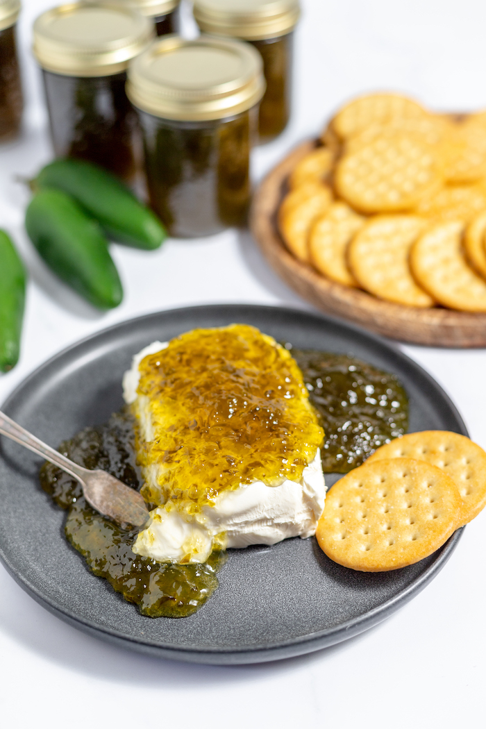 Image shows jalapeno jelly poured over top of cream cheese on a plate. Crackers sit on a plate nearby. 