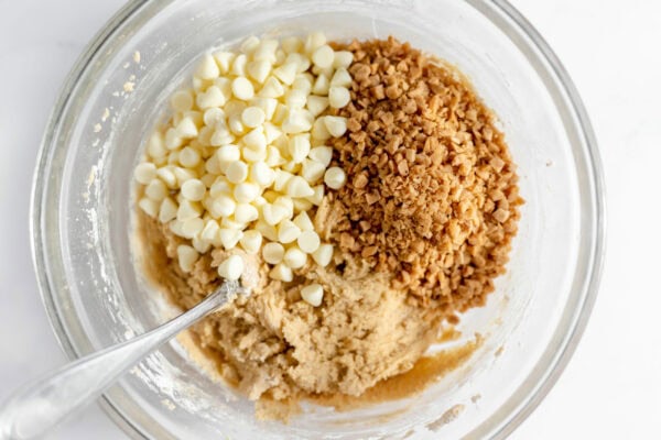 Image, taken from above, shows a glass bowl with dough to make espresso toffee cookies, as well as white chocolate and toffee bits to mix in.