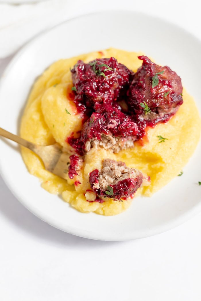 Image shows a serving of cranberry meatballs served over polenta on a white plate with a fork in it. 