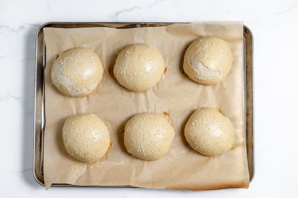 Image, taken from above, shows baked sourdough hamburger buns on a cookie sheet and sprinkled with sesame seeds