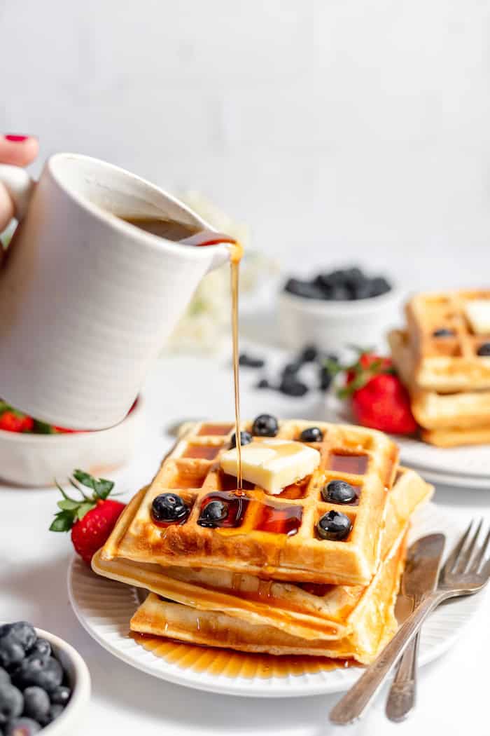 Image shows a hand pouring syrup over sourdough waffles topped with butter and blueberries on a plate.