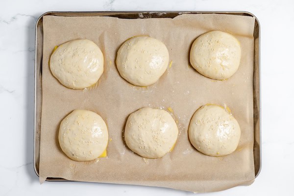Image, taken from above, shows sourdough hamburger buns with sesame seeds ready to bake on a cookie sheet