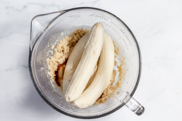 Image, taken from above, shows three bananas being added to the sourdough dough in a large glass mixing bowl